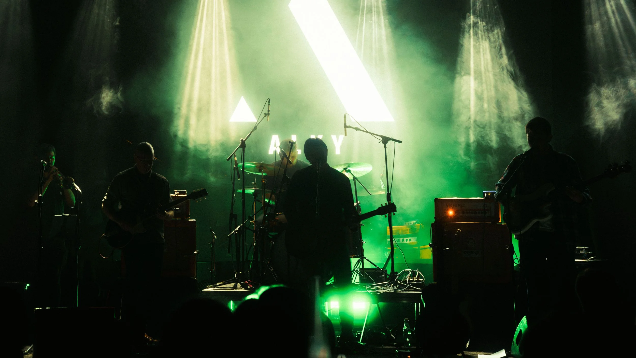 A band performs on stage with green lighting, silhouettes of five musicians playing instruments, and a large geometric logo with the letters 'AKY' in the background.