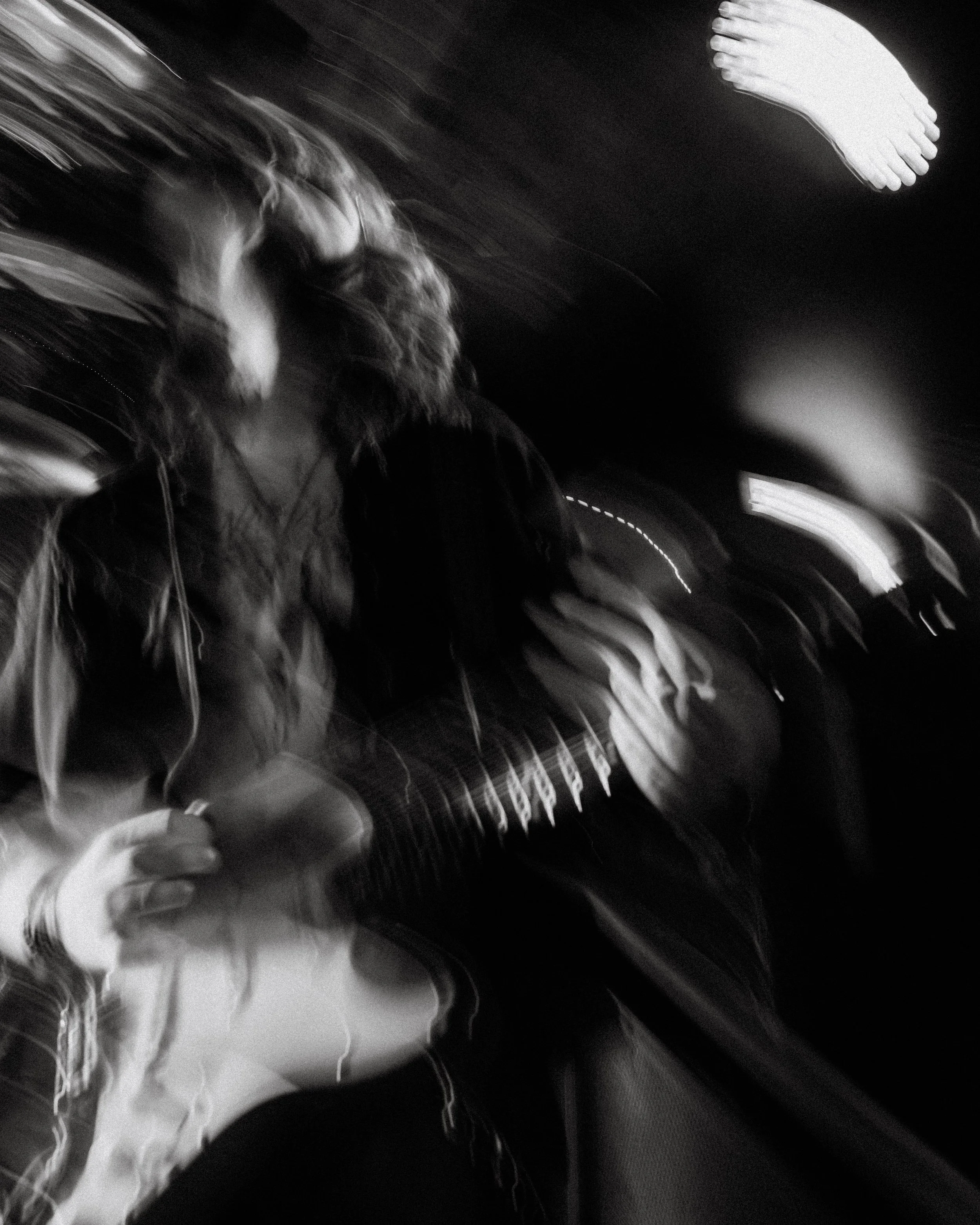 Blurred black and white photo of a person with long hair holding a guitar, sitting in a car at night with streaks of light from the moving vehicle.