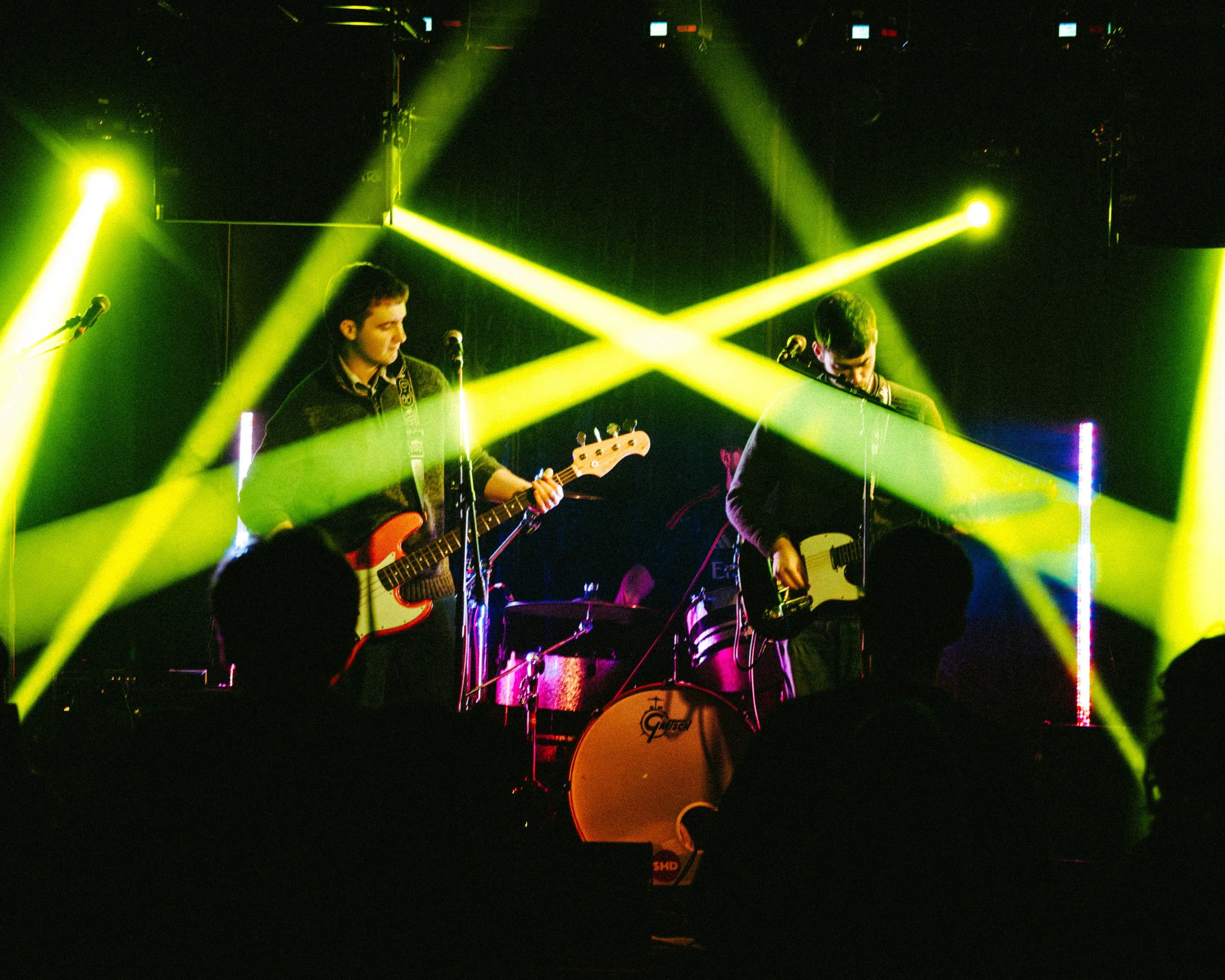 Two musicians performing on stage with yellow stage lights, each playing an electric guitar, and a drummer in the background.
