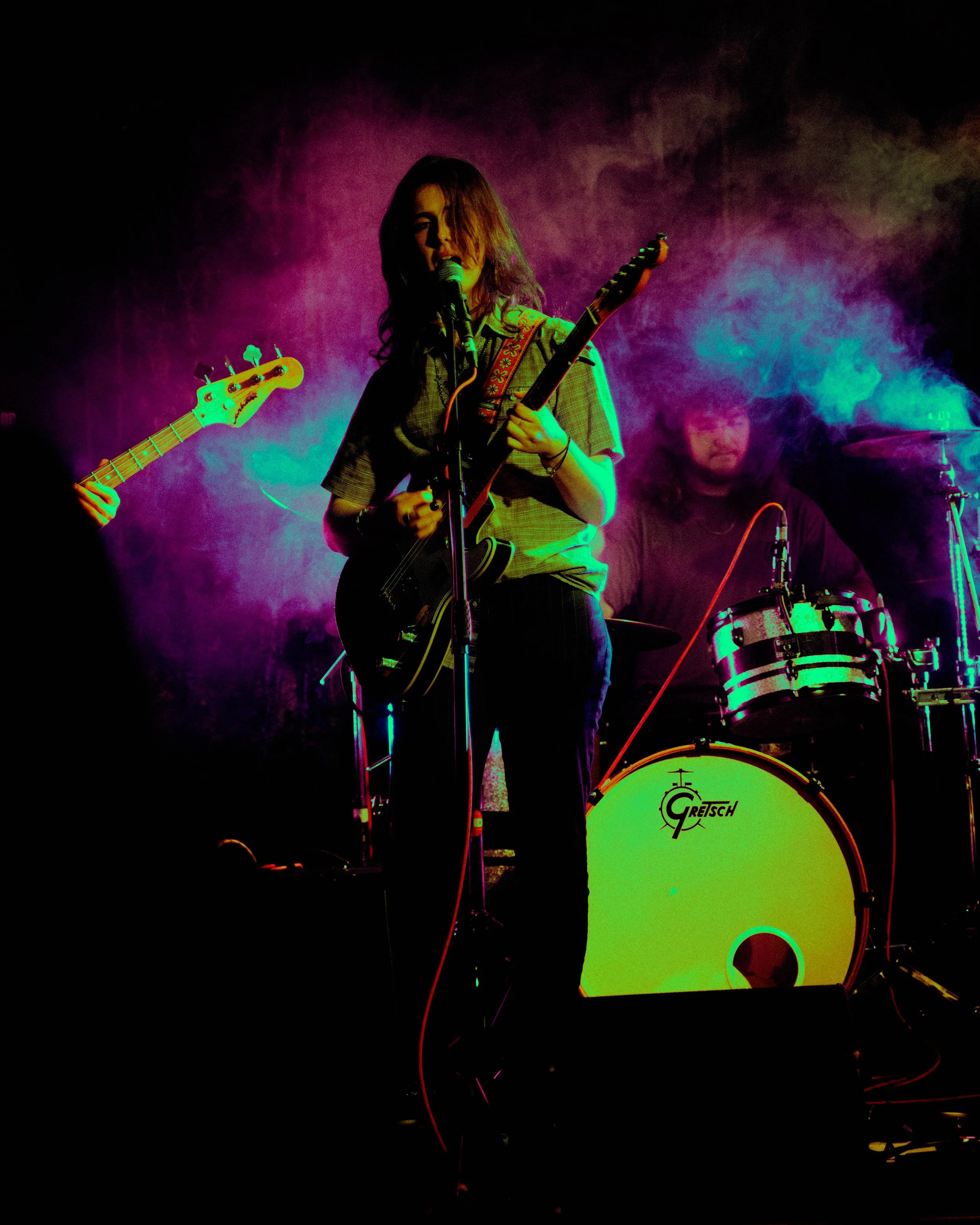 Female musician playing guitar and singing into a microphone with a drummer in the background on stage, colorful lighting and smoke effects.