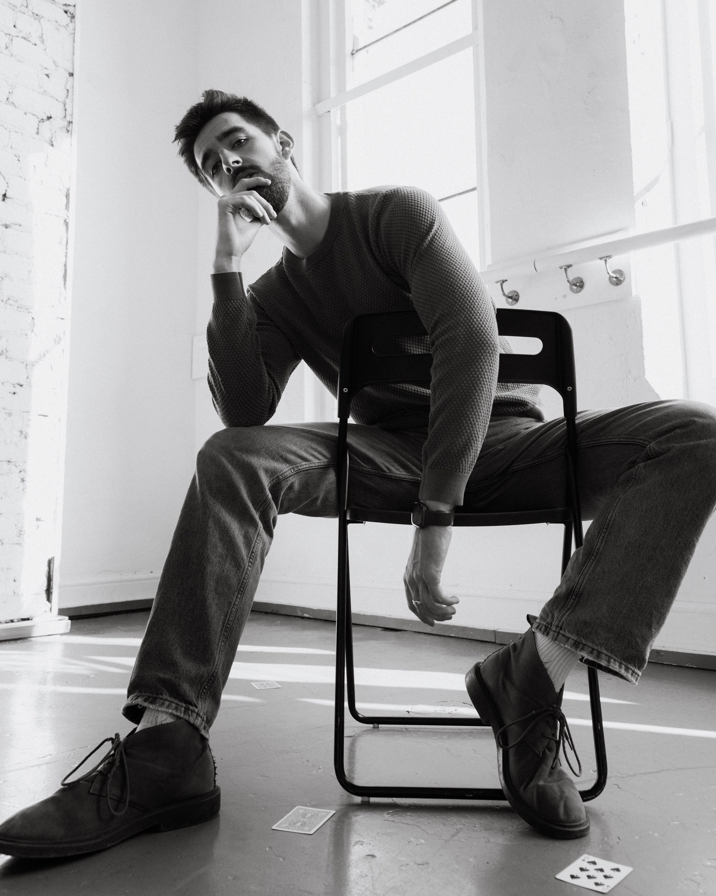 Black and white photo of a man sitting on a chair in a room with large windows. He has a beard and mustache, wearing a checkered sweater, jeans, and boots, with a watch on his wrist. His head is tilted, resting on his hand, and he is looking at the c