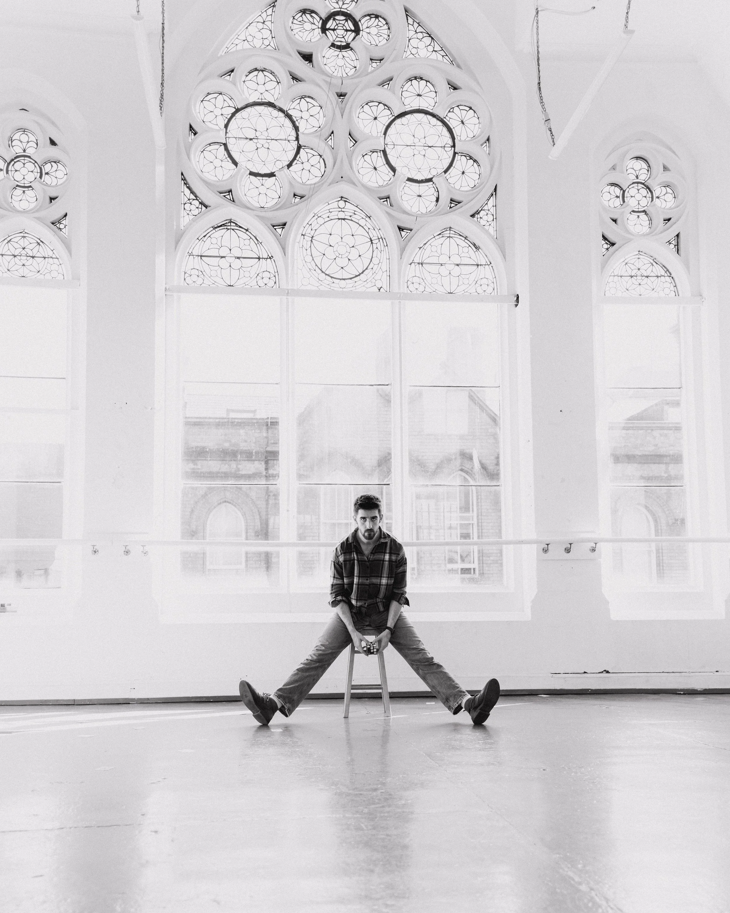 A young man with a beard and plaid shirt sitting on a chair with his legs spread wide open, holding a camera. The background features large arched windows with intricate stained glass designs, within a spacious room.