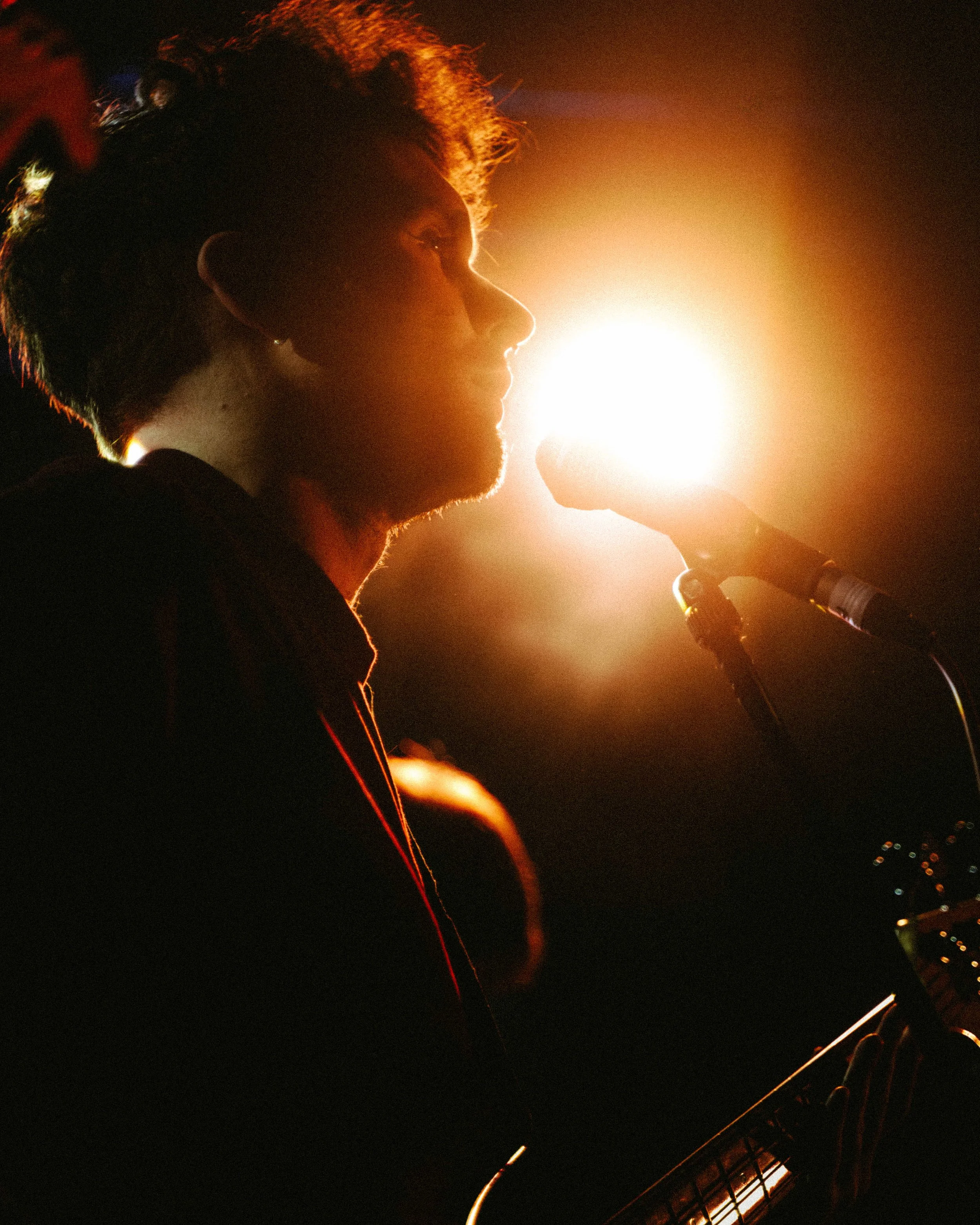 A silhouette of a person singing or speaking into a microphone, with bright stage lights behind them, creating a glowing effect around their profile.
