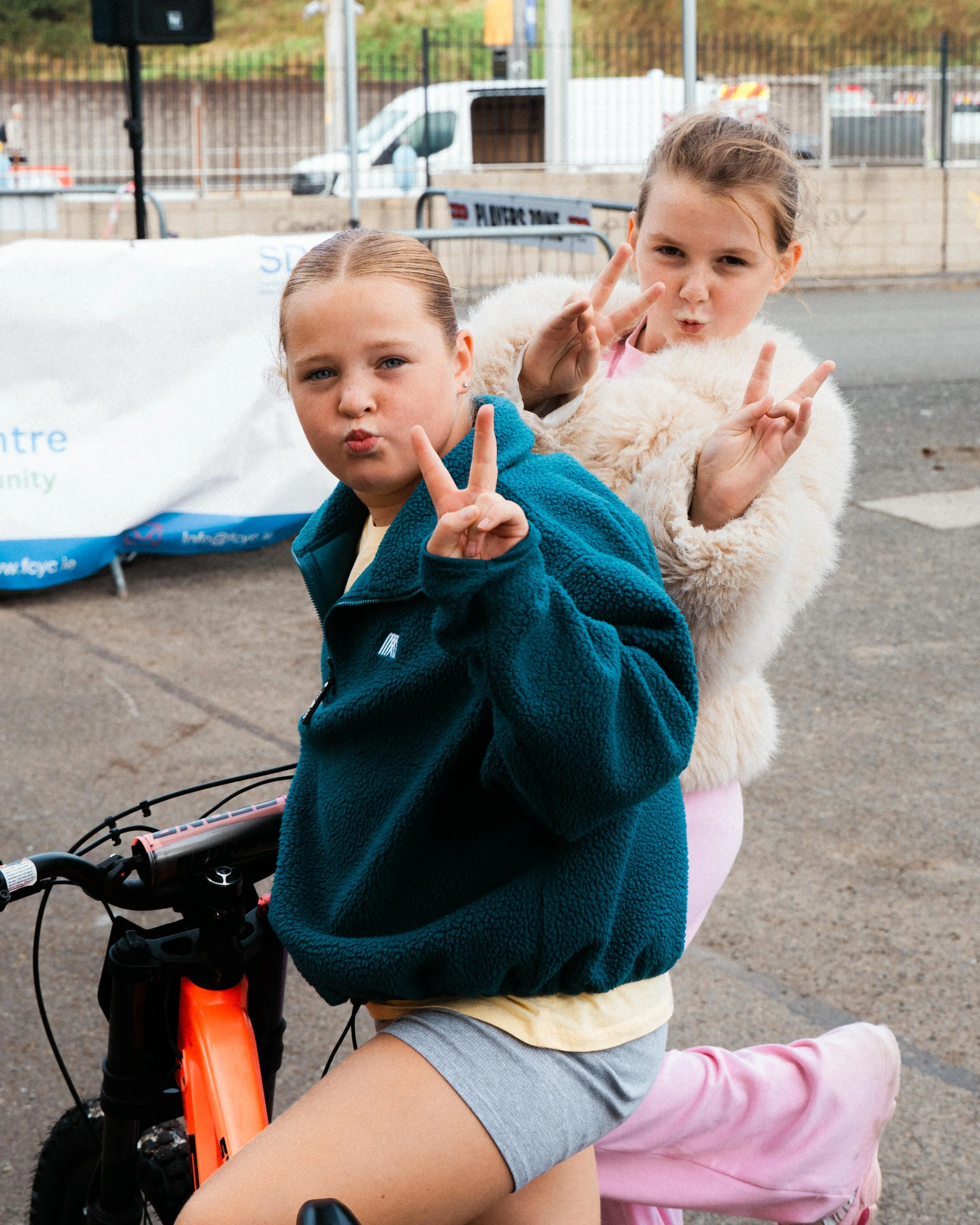 Two young girls making peace signs and puckering lips while sitting on a sidewalk, with a bicycle and a white van in the background.