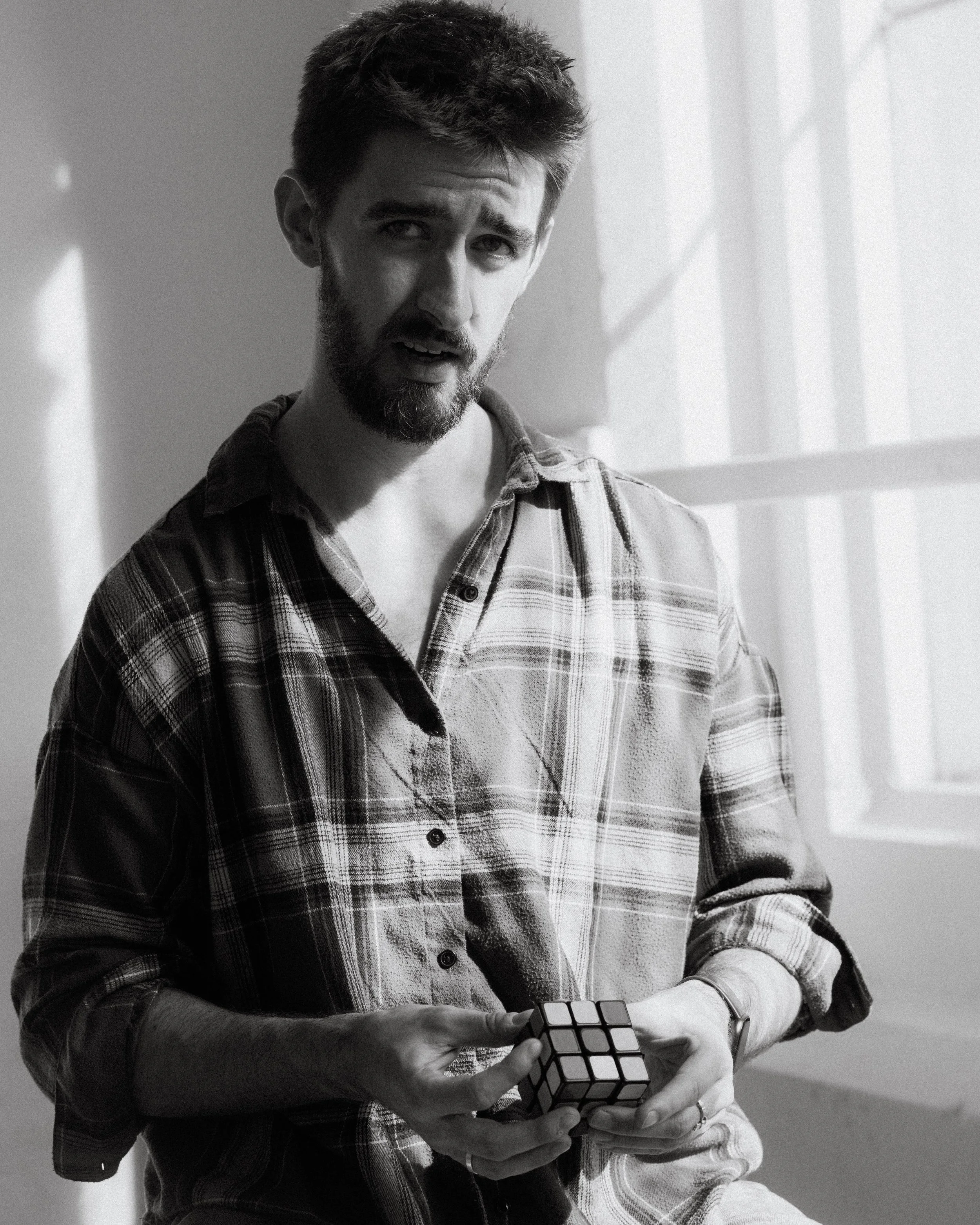 A young man with a beard and short, messy hair holding and looking at a Rubik's cube. He is wearing a plaid shirt and a watch, sitting indoors near a window with natural light.