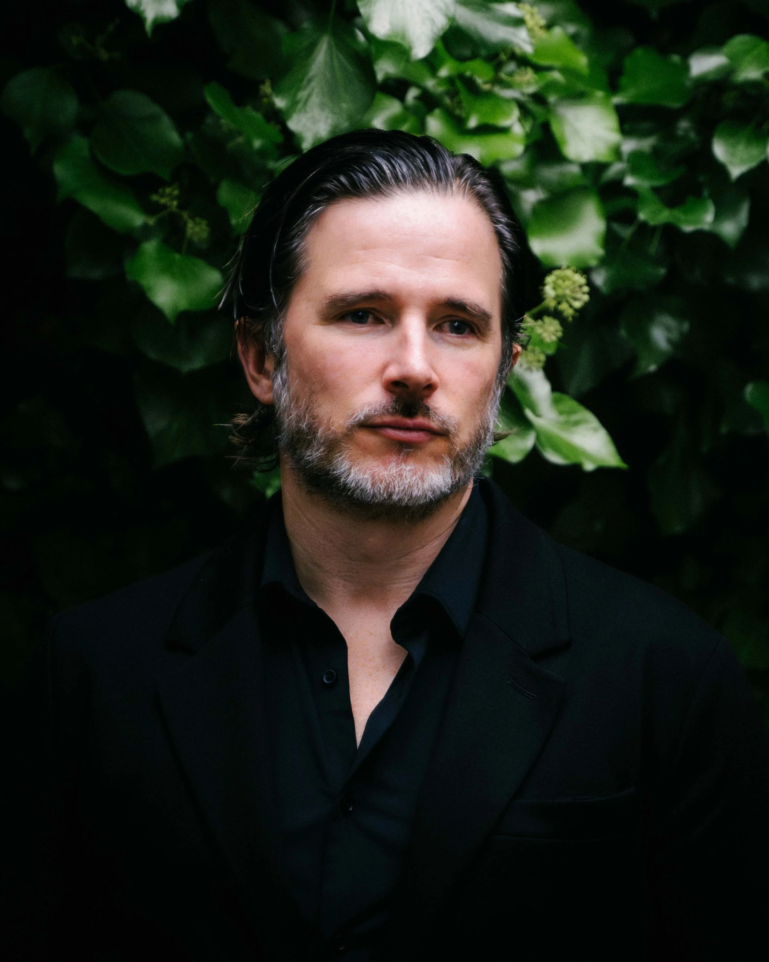 Head and shoulders of a man with dark, slicked-back hair and a beard, wearing a black shirt, against a background of green foliage.