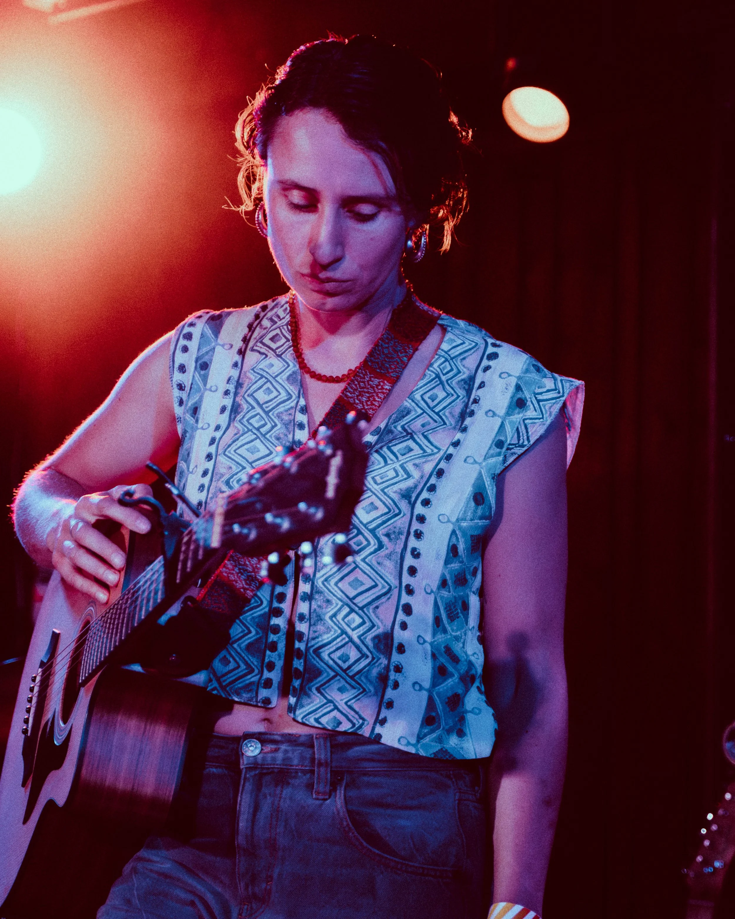 A woman with short dark hair wearing a patterned sleeveless top, playing an acoustic guitar on stage with red stage lighting.