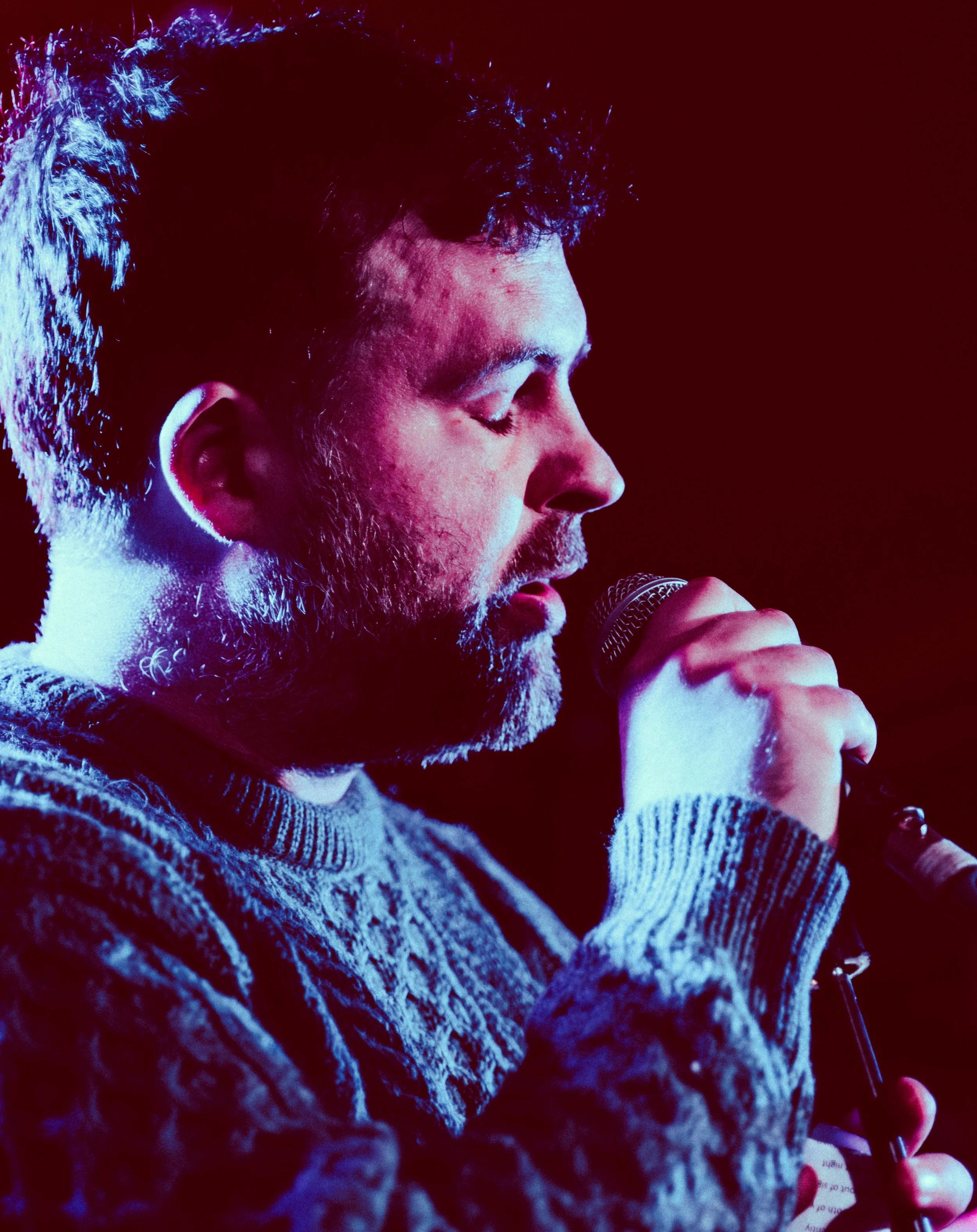 Close-up of a man with a beard, closed eyes, wearing a patterned sweater, holding a microphone, with a dark red background.