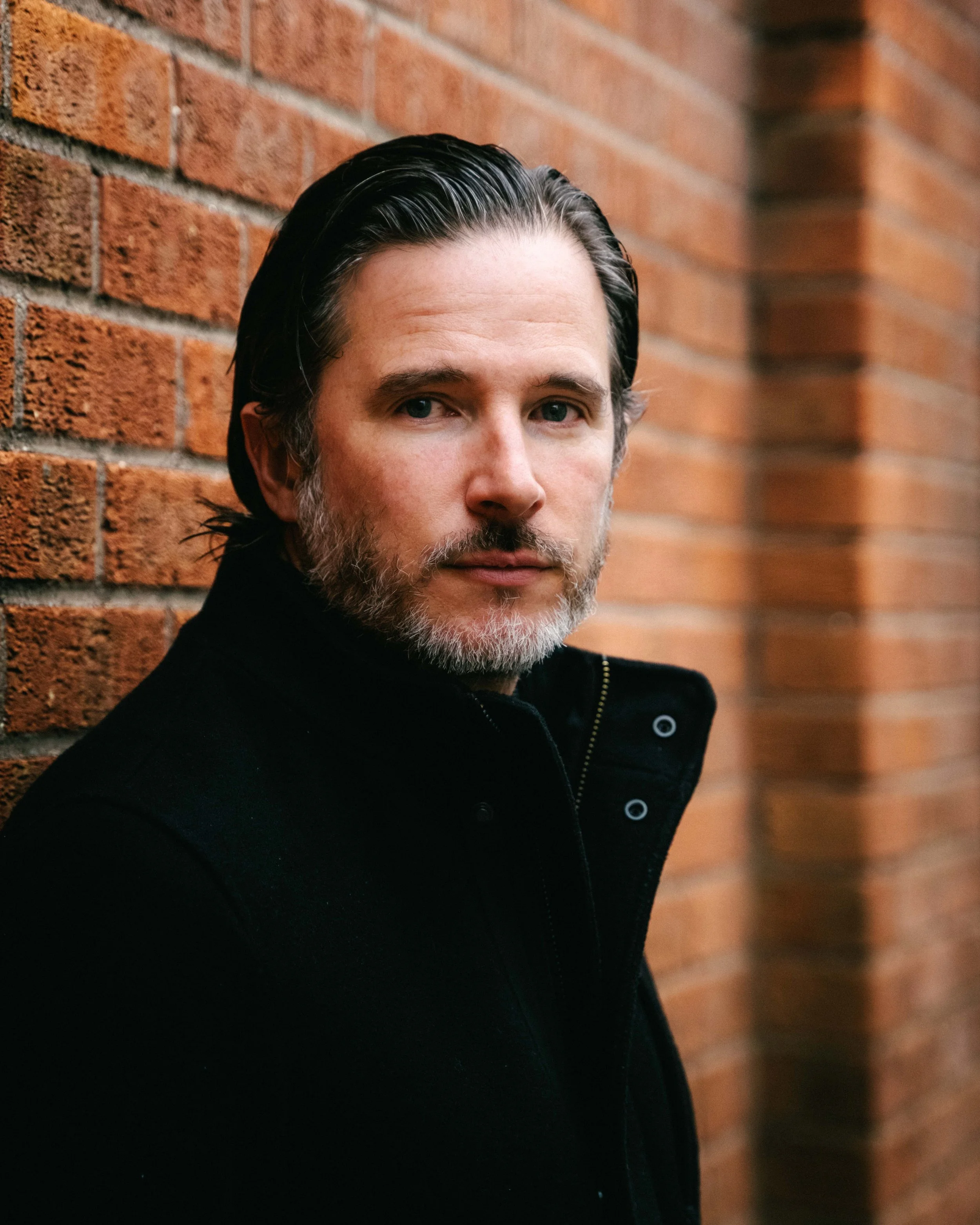 Close-up of a middle-aged man with slicked-back dark hair and a beard, standing against a red brick wall, wearing a black jacket.