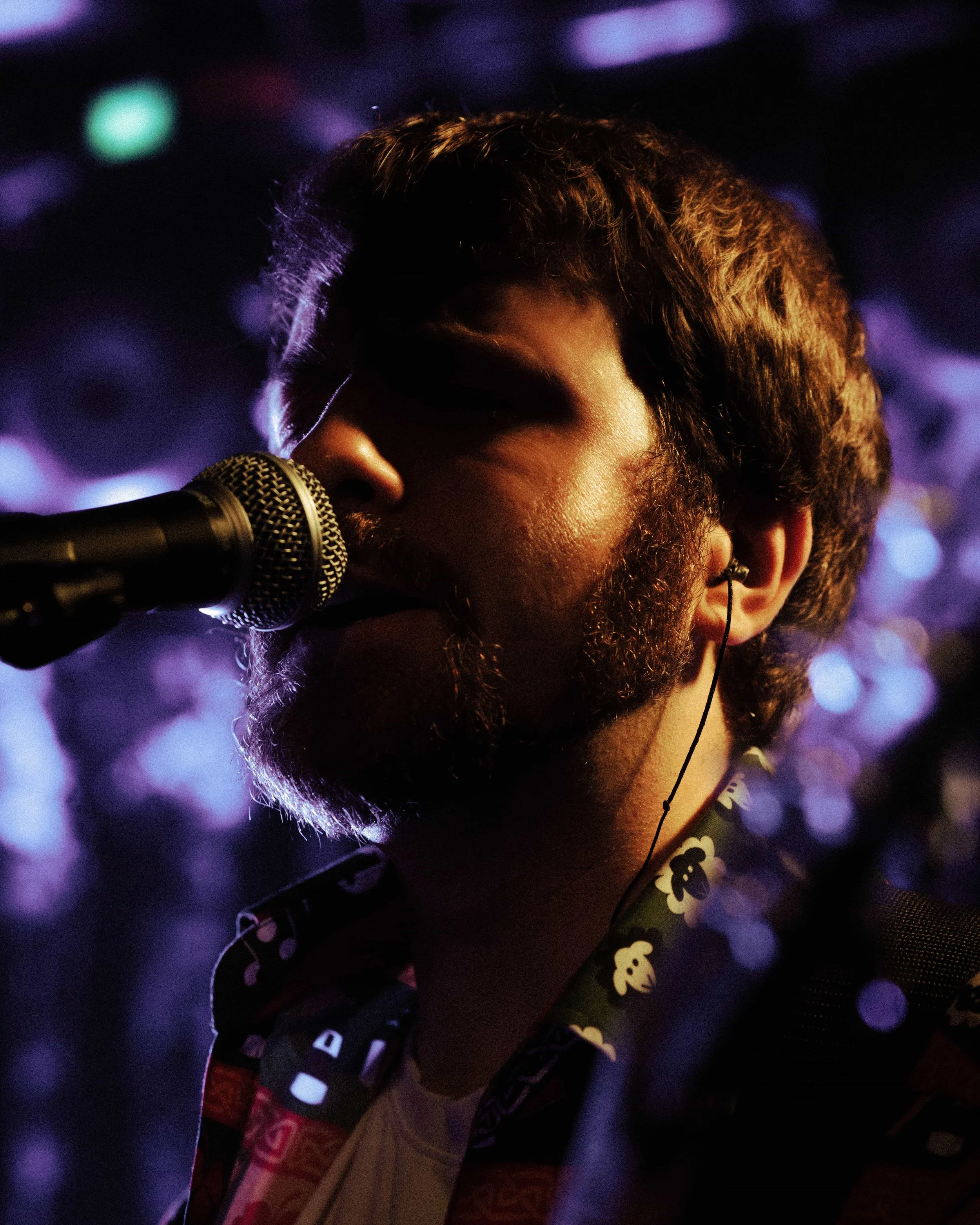 A man with a beard singing into a microphone at night with blurred purple and blue lights in the background.