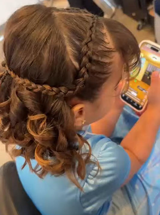 A young girl with styled braided hair and curly ends, wearing a light blue shirt, sitting and looking at her phone.