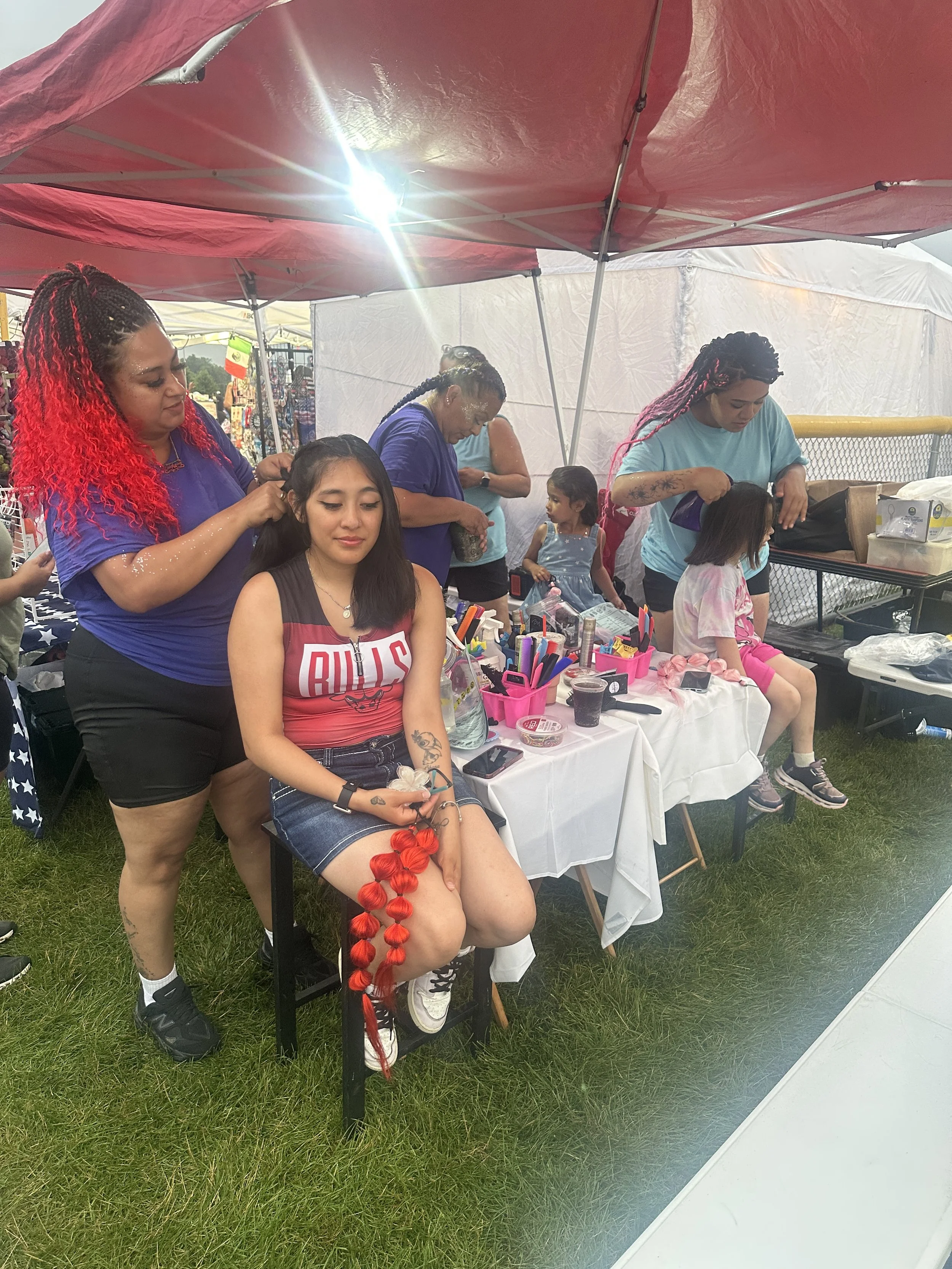 A woman with bright red hair styles the hair of a seated young girl with black hair. Two women are working behind a table with beauty supplies at an outdoor event under a red canopy.
