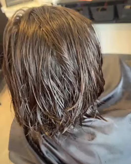 Person with wet, shoulder-length brown hair sitting in a salon chair, receiving a haircut or hair treatment.