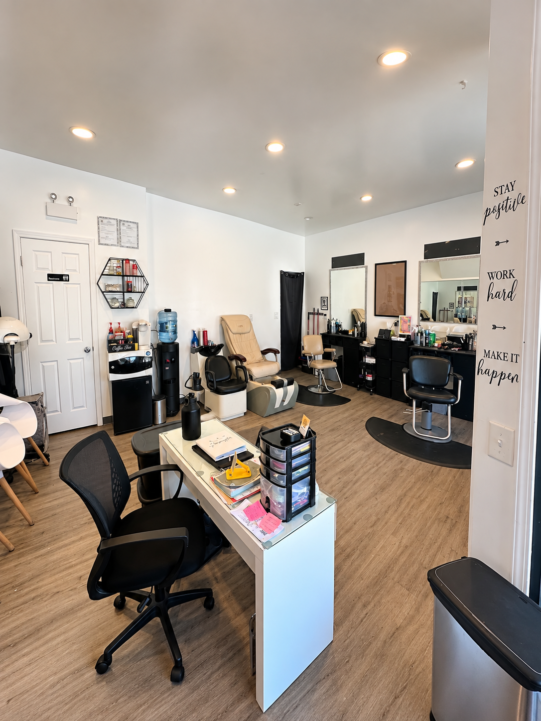 Interior of a nail salon with manicure stations, a desk with office supplies, massage chairs, and shelves with nail and salon products, all in a bright, clean space.