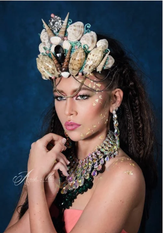 Woman wearing a headdress decorated with seashells, coral, and other marine elements, and elaborate jewelry with gemstones, against a dark blue background.