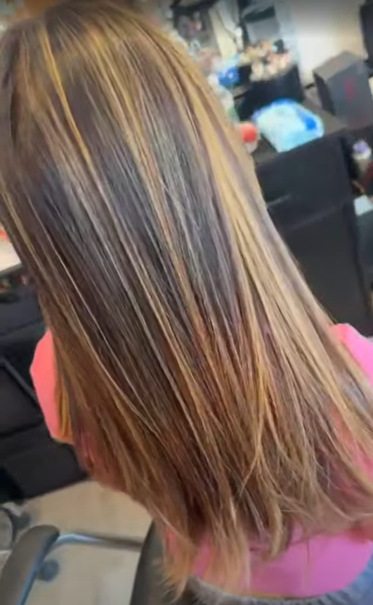 Back of a woman's head with long, straight, shiny brown hair with highlights, inside a hair salon.