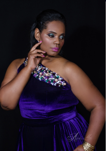 Woman in a purple velvet dress with jeweled embellishments on the shoulder, posing with her hand near her face against a black background.
