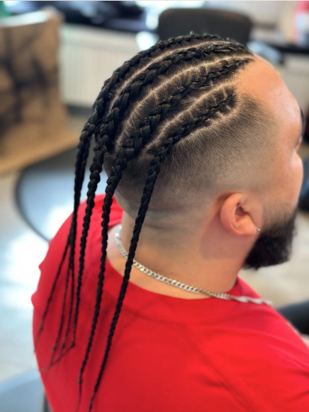 Man with cornrows and a fade haircut, wearing a red shirt and a silver chain, sitting in a barbershop.