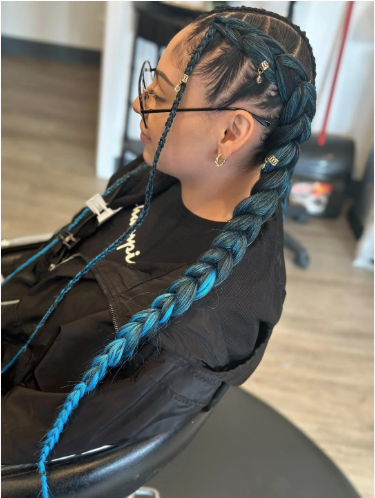 Side profile of a woman with braided hair dyed blue, wearing glasses and gold jewelry, sitting in a salon chair.