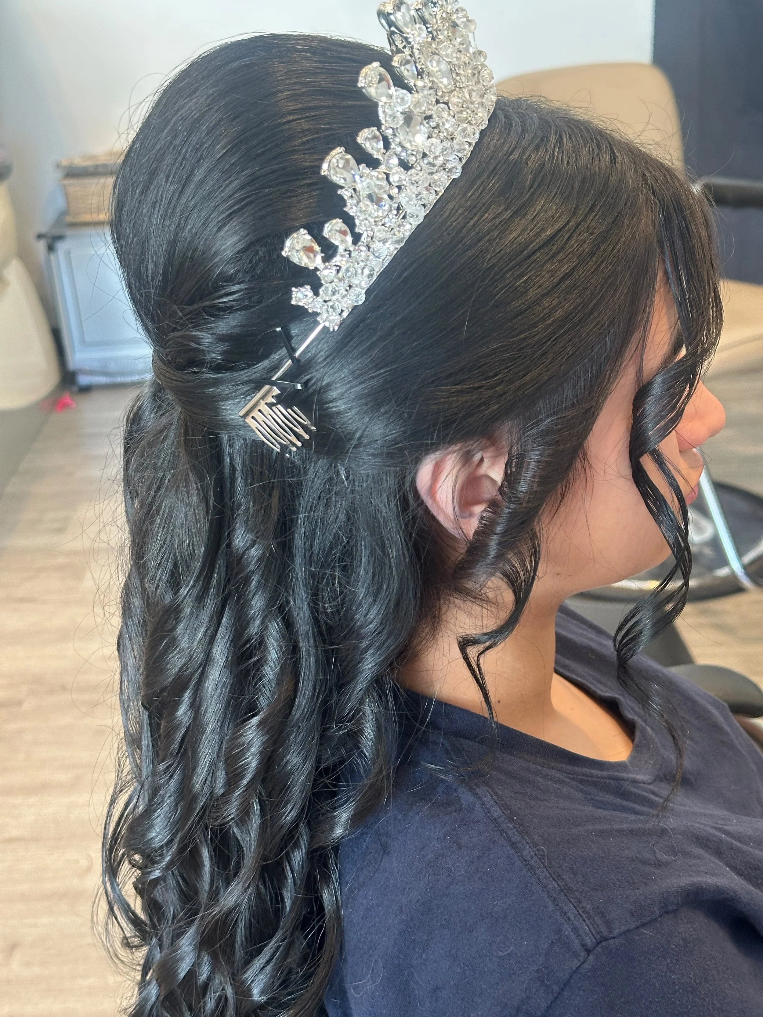 A woman with black hair styled in loose curls, wearing a silver tiara with clear gemstones, and a black top, sitting in a salon chair.
