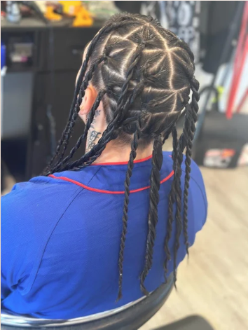 Person with styled cornrow braids sitting in a salon chair, wearing a blue shirt with red trim.