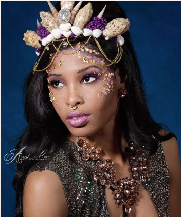 Woman wearing a large decorative headpiece with shells, crystals, and purple accents, gold chains, and glitter makeup, posing against a dark background.