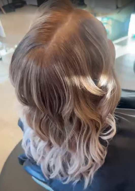 A person's head with styled, wavy, ombre hair color that transitions from light brown at the roots to platinum blonde at the ends.