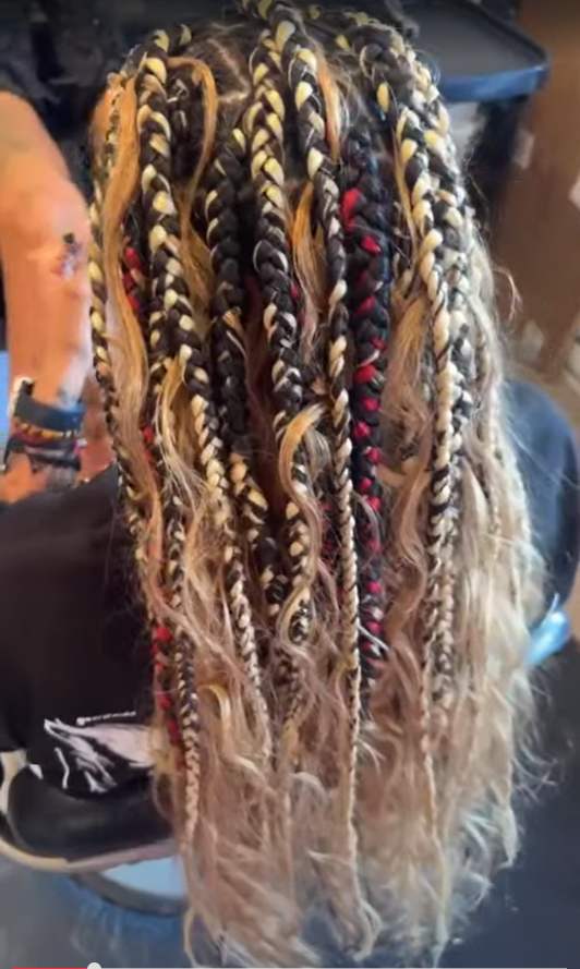 Close-up of a woman's head with long, intricately styled braids with blonde, black, and red accents, inside a salon.