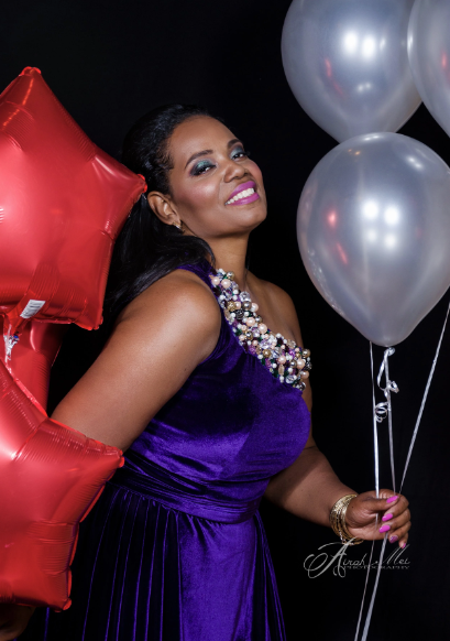 Woman in purple dress holding silver balloons, with red star-shaped balloons beside her, smiling, against a black background.