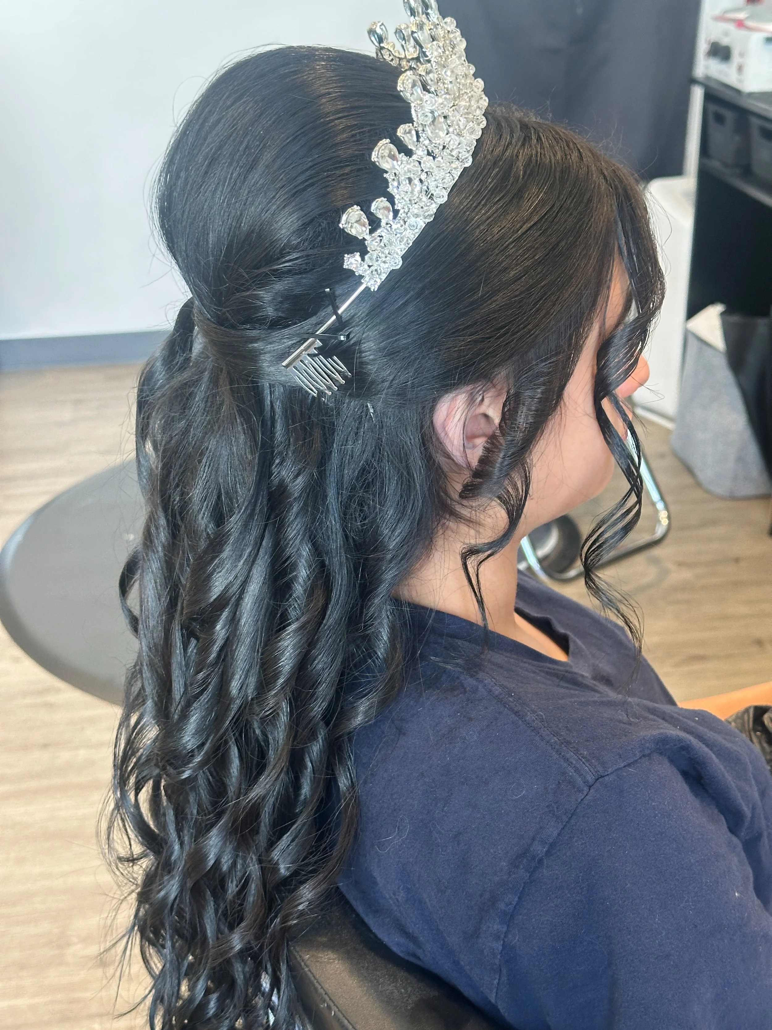 Woman with dark, wavy hair styled in loose curls, wearing a sparkly tiara and black hair clip, sitting in a salon chair.
