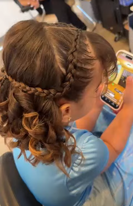 A woman with intricately braided and curled hair, wearing a light blue shirt, seated and looking at her phone in a setting that appears to be a salon or similar environment.
