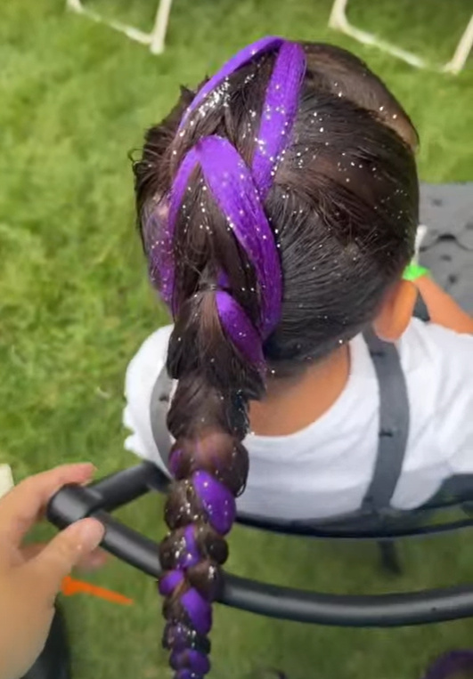 Child with braided hair dyed purple, sitting outdoors on a black chair on a grassy area, with sparkle glitter in hair, wearing a white shirt.