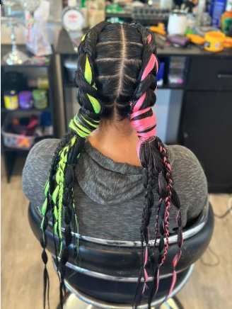 A person with long braided hair with sections dyed neon green and pink, seated in a salon chair with black shelves and hair styling tools in the background.