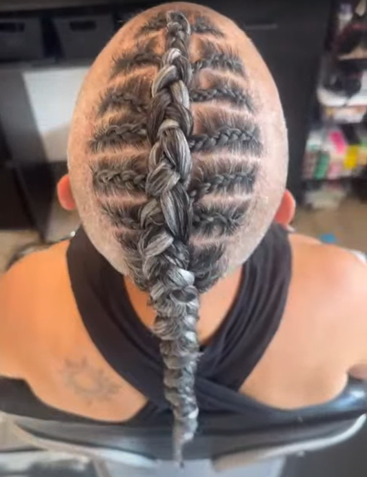 Back of a head with a unique braided hairstyle: a large central braid with skull motifs shaved into the scalp and smaller braids around it.