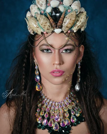 A woman with elaborate seashell and jewelry headdress, wearing large colorful gemstone necklace and earrings, with makeup and long dark curly hair against a dark background.