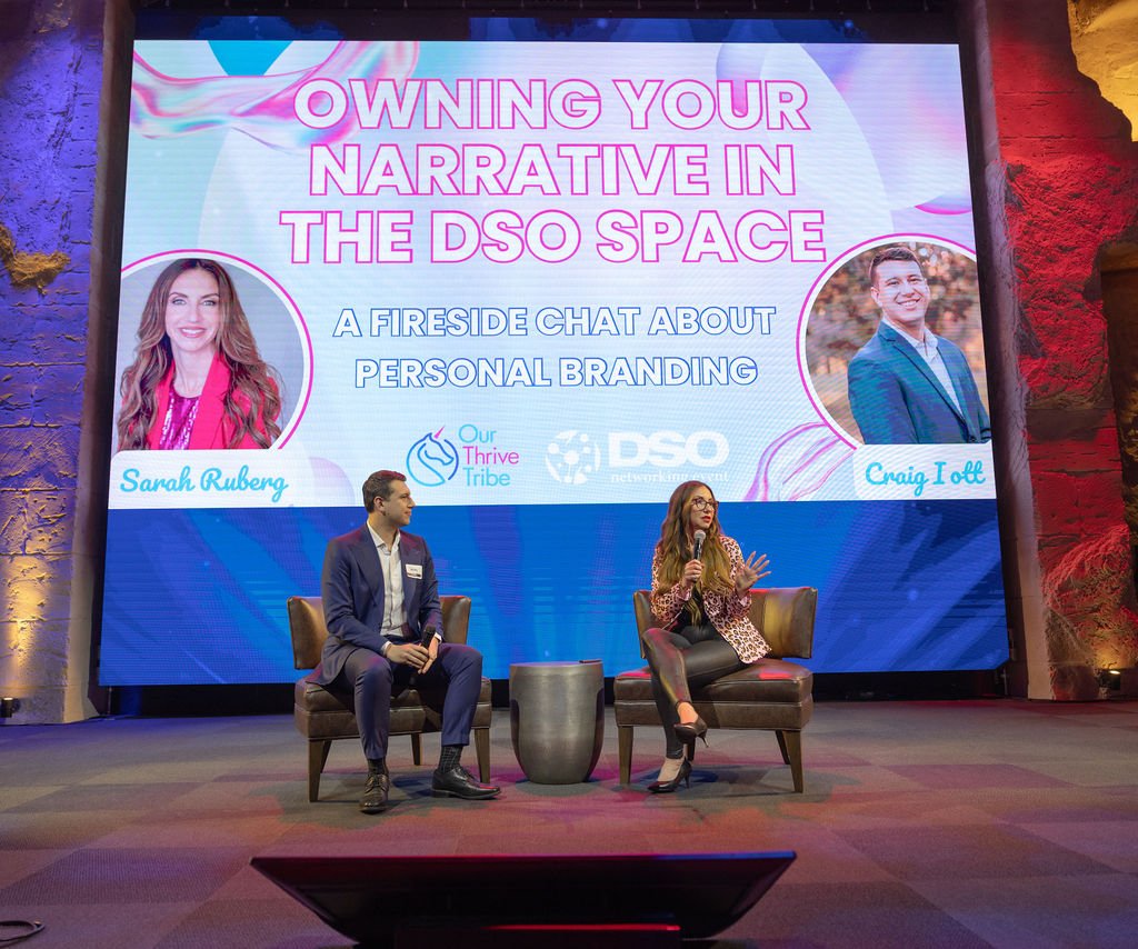Panel discussion with two speakers, a man and a woman, seated on stage in front of a large screen displaying information about a fireside chat titled "Owning Your Narrative in the DSO Space." The screen features photos of Sarah Ruberg and Craig Iott,