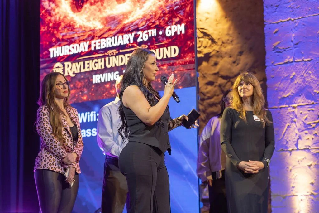 Woman speaking into a microphone on stage during an event, with three women standing behind her and a large screen displaying event details in the background.