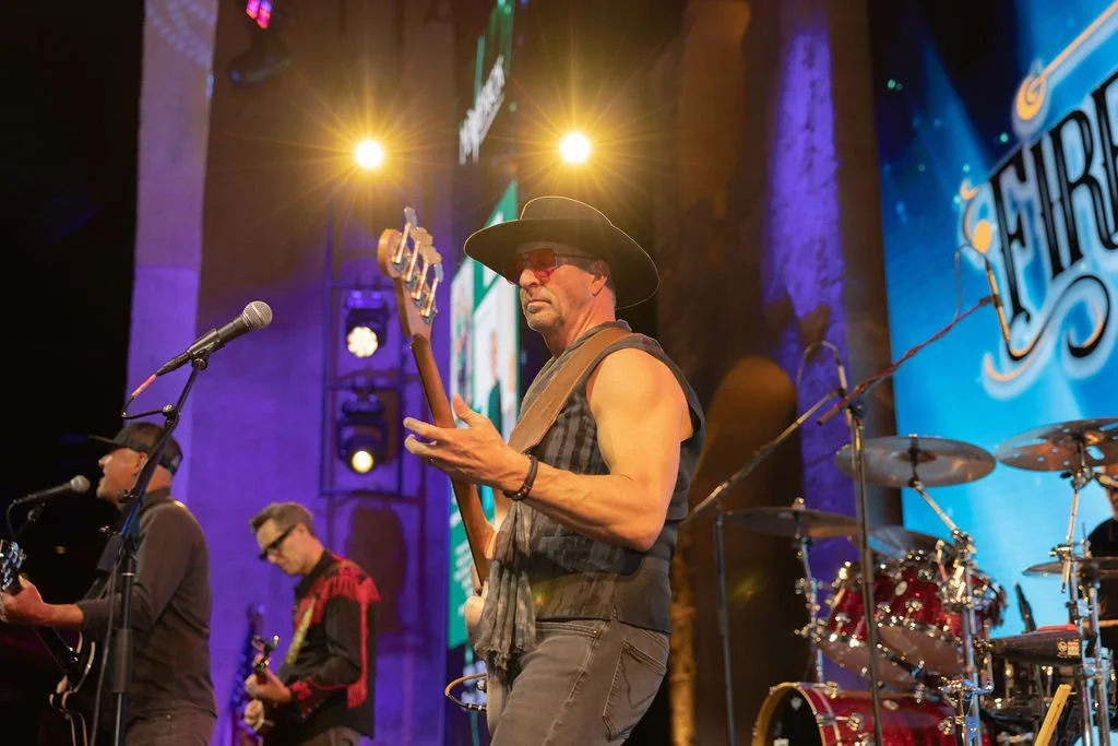 Musicians performing on a stage with colorful lighting, including a man playing guitar in the foreground wearing a black cowboy hat, sunglasses, and a sleeveless shirt, with a drum set and other band members in the background.