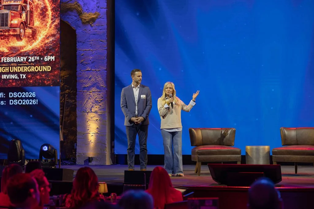 People on stage at a conference or event, with a woman speaking into a microphone and a man standing beside her, in front of a large blue screen and chairs, with an audience seated below.