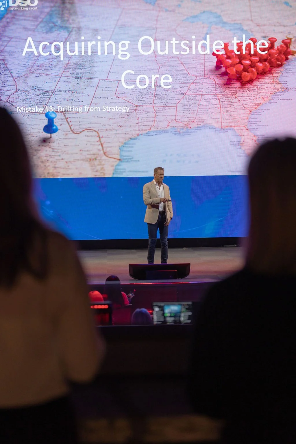 A speaker stands on stage in front of a large screen displaying a map of the United States with pins. The presentation slide reads 'Acquiring Outside the Core' with a subtitle 'Mistake #3: Drifting from Strategy'. Audience members are visible in the 