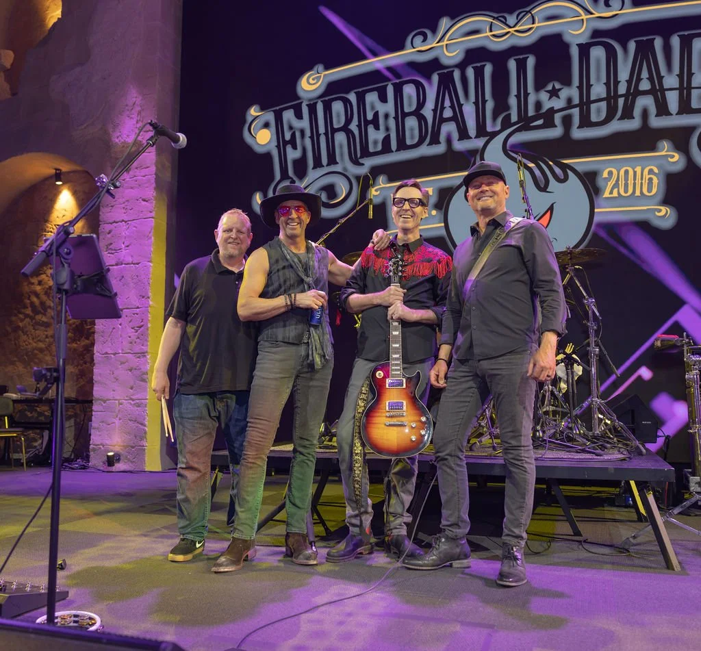 Four male musicians standing on stage with musical instruments, smiling at the camera, at the Fireball Daze event in 2016.