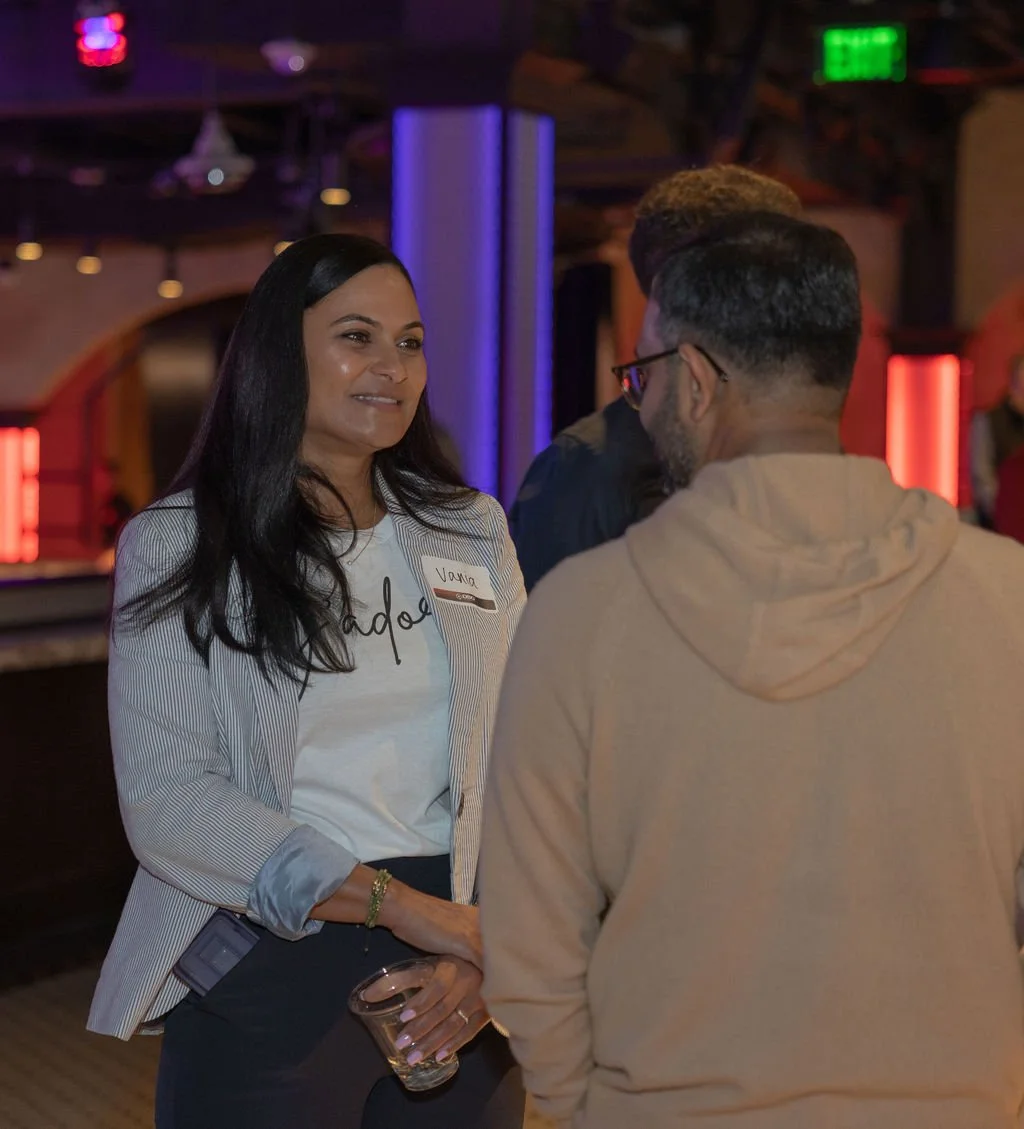 A woman with long dark hair smiling and holding a drink in one hand, engaged in conversation with a man wearing glasses and a beige hoodie, in a dimly lit bar or club setting with colorful lighting.