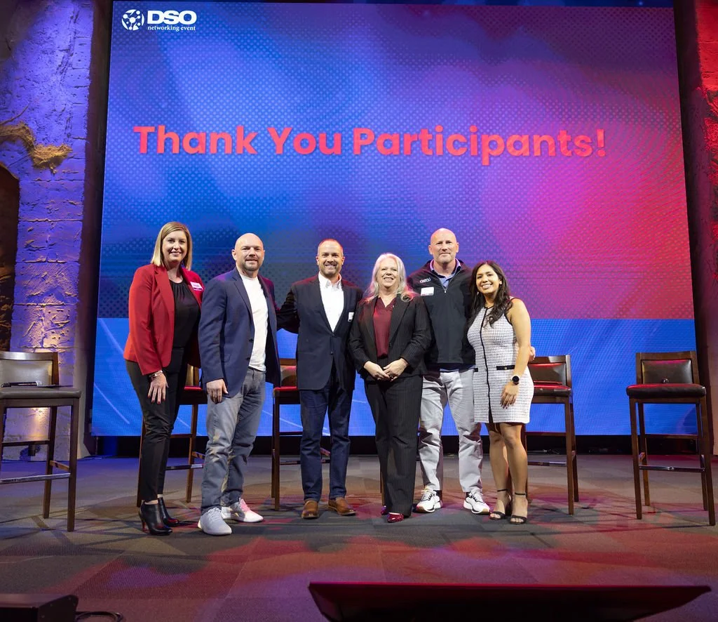 Group of six people standing in front of a large screen displaying 'Thank You Participants!' at a networking event, with chairs on stage.