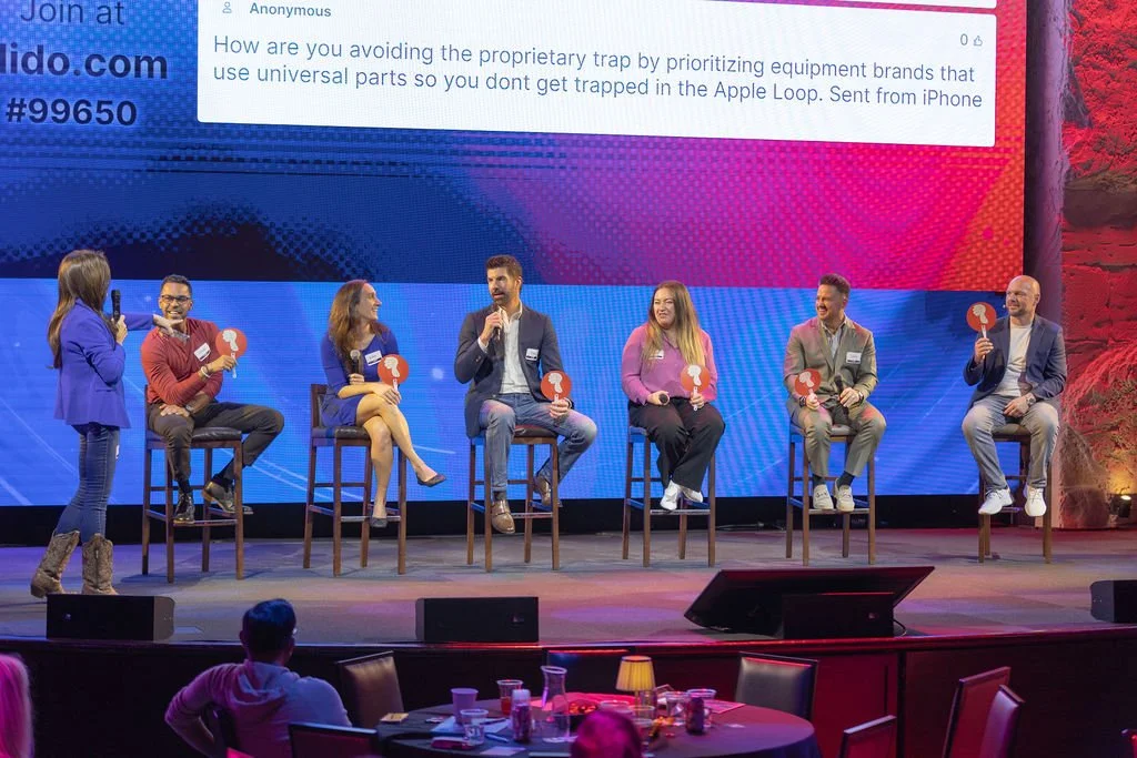 Panel discussion with seven people on stage, five sitting on stools, two standing, holding icons, in front of a large screen displaying text, audience in foreground.