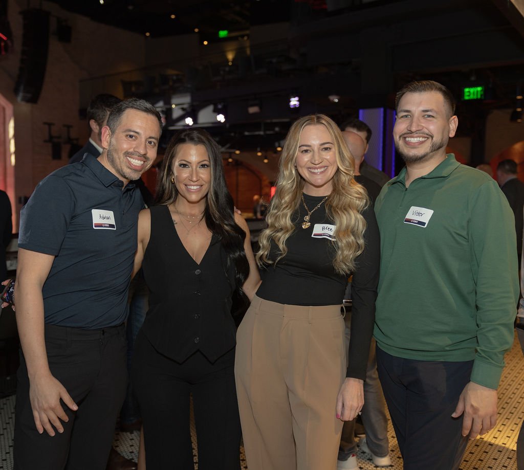 Four people standing together at a social event, smiling at the camera. They are wearing casual to business casual attire, with name tags on their shirts.