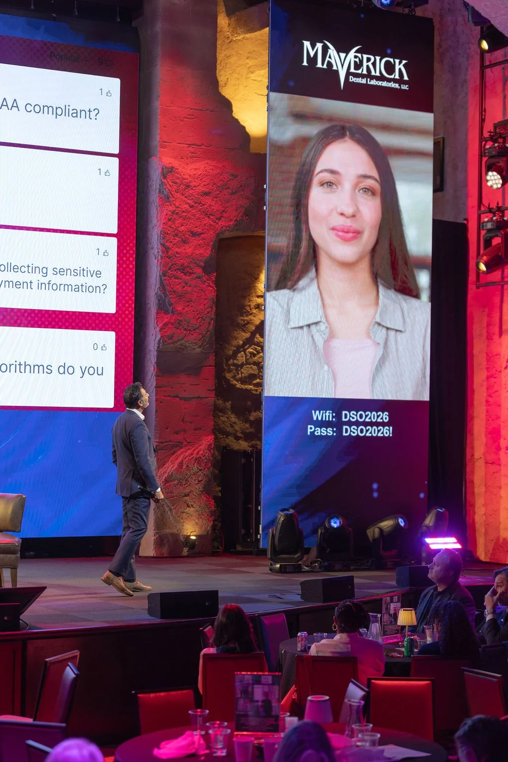 Large digital screen showing a woman with long brown hair smiling, with WiFi and password details; the logo for Maverick Dental Laboratories at the top; a conference stage with a man in a suit standing and looking up at the screen; audience members s