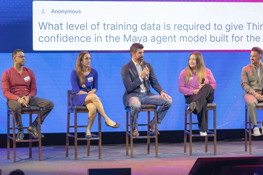 A panel of five people sitting on chairs on a stage, participating in a discussion or Q&A session, with a large screen behind them displaying a question about training data for a Maya agent model.