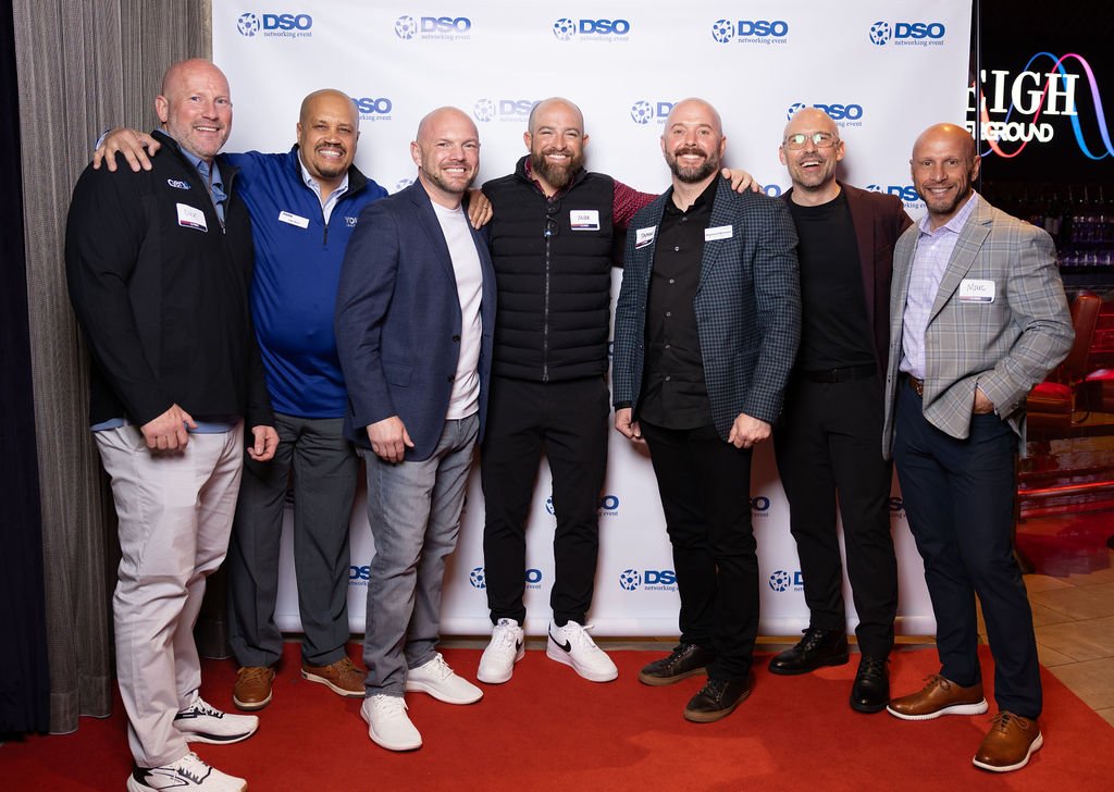 Group of seven men smiling and standing together at an event with a backdrop featuring the DSO logo, dressed in business casual attire.