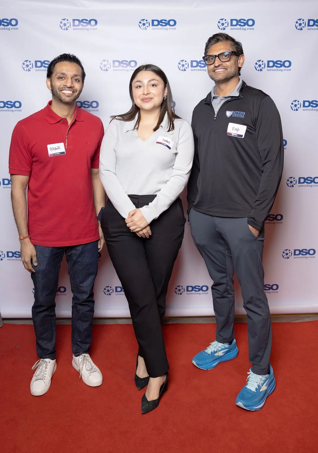 Three people standing in front of a backdrop with the DSO logo at a networking event. The person on the left is a man wearing a red polo shirt and jeans, smiling. The person in the middle is a woman in a white blouse and black pants, also smiling. Th
