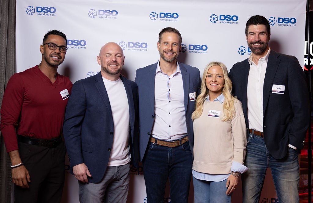 Five people standing together at a networking event in front of a banner with the DSO logo. They are smiling and dressed in business casual attire, four men and one woman.